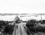 Military vehicles move ashore from Mulberry Artificial Harbor A, across a pontoon bridge, to Omaha Beach, Normandy, France, June 16 1944 (D+10). Visible are M3 Halftracks, an M8 Greyhound, and AFKWX 2½-ton Trucks.