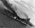 Angled view of the carrier Franklin burning and listing badly after bomb hits aft set off more bombs and fueled aircraft, 19 Mar 1945. Note burning fuel pouring off the hangar deck.