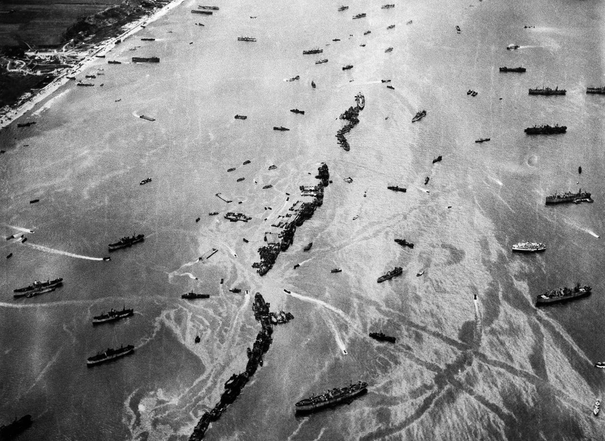 13 Liberty Ships deliberately scuttled to form a breakwater for invasion vessels landing on the Normandy beachhead lie in line off the beach shielding the ships in shore.