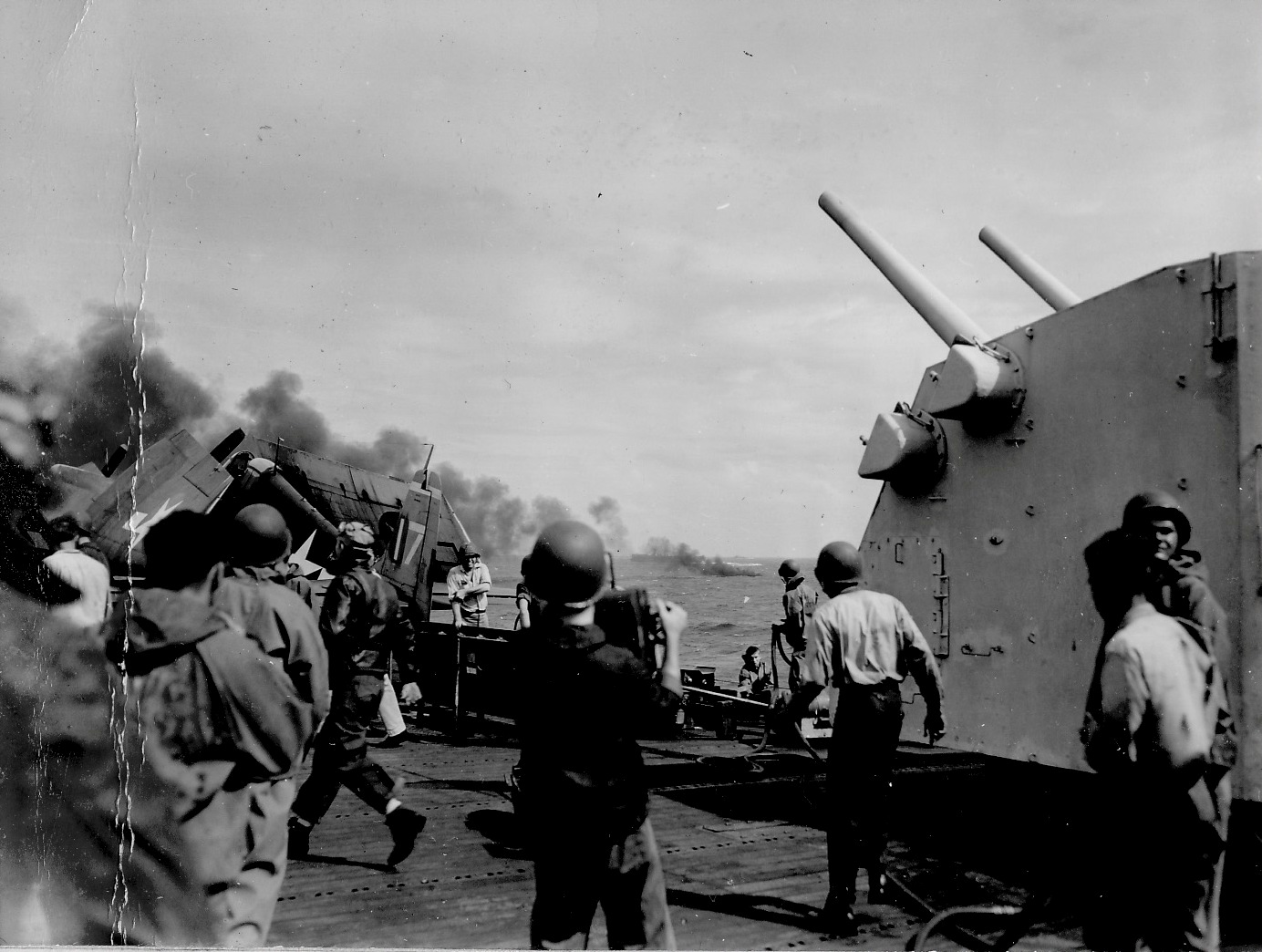 Activity continues on the flight deck of USS Essex as a crashed Japanese airplane burns off the starboard bow, 24 Oct 1944 off Luzon in the Philippines. Note Number 1 5-inch/38 caliber gun mount at right.