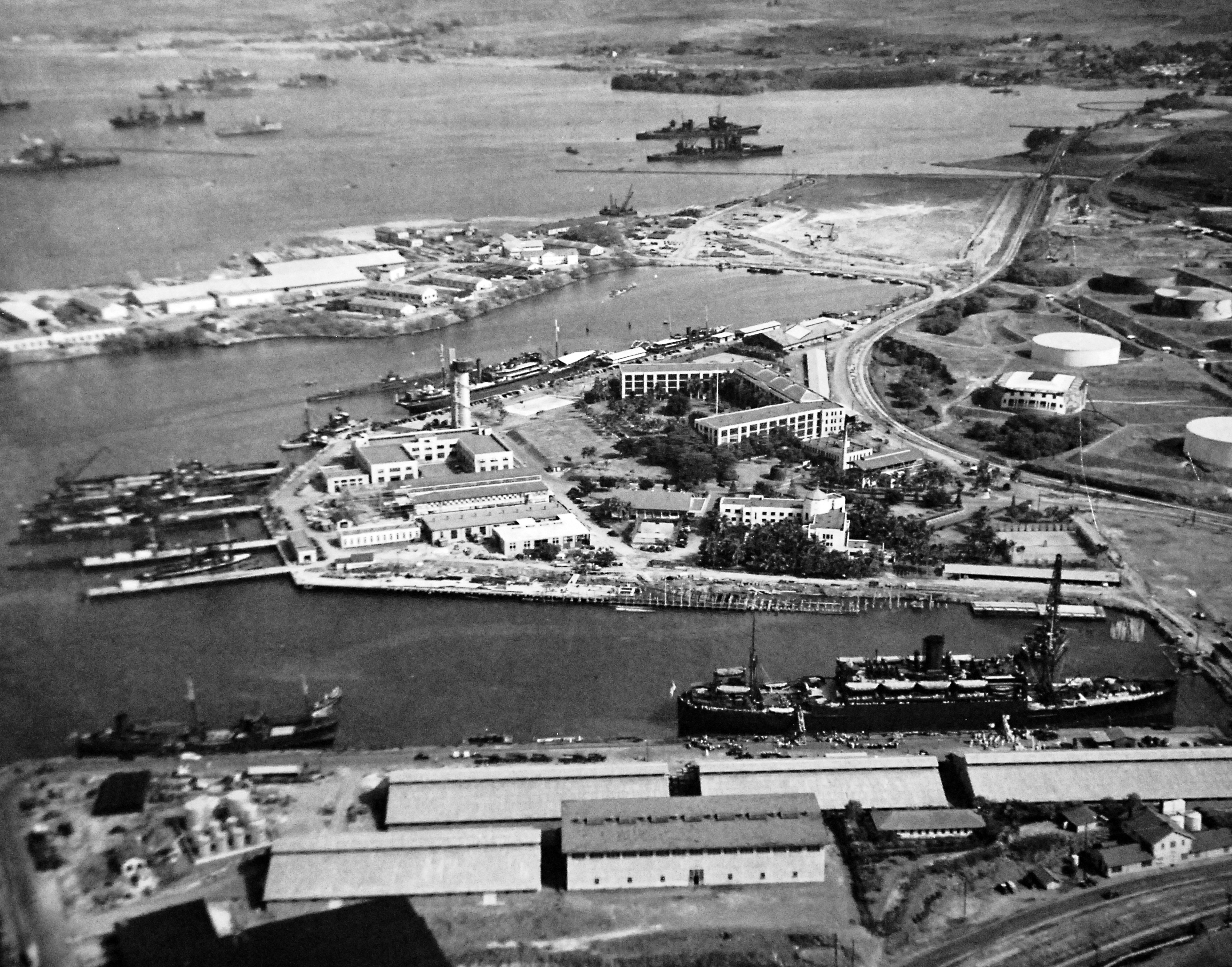 Aerial view of the Pearl Harbor Submarine Piers at Merry Point, 13 Oct 1941. Also pictured is the U-shaped Headquarters Building for the Pacific Fleet and a tank farm at right. Note one tank painted as a building.