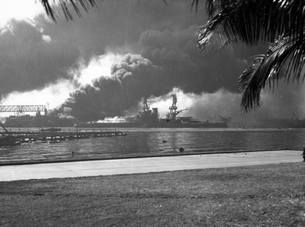 USS Nevada, down by the head, beached at Hospital Point in Pearl Harbor, Hawaii during the Japanese air attack on 7 Dec 1941. Note smoke from destroyer USS Shaw in the floating drydock just beyond Nevada.