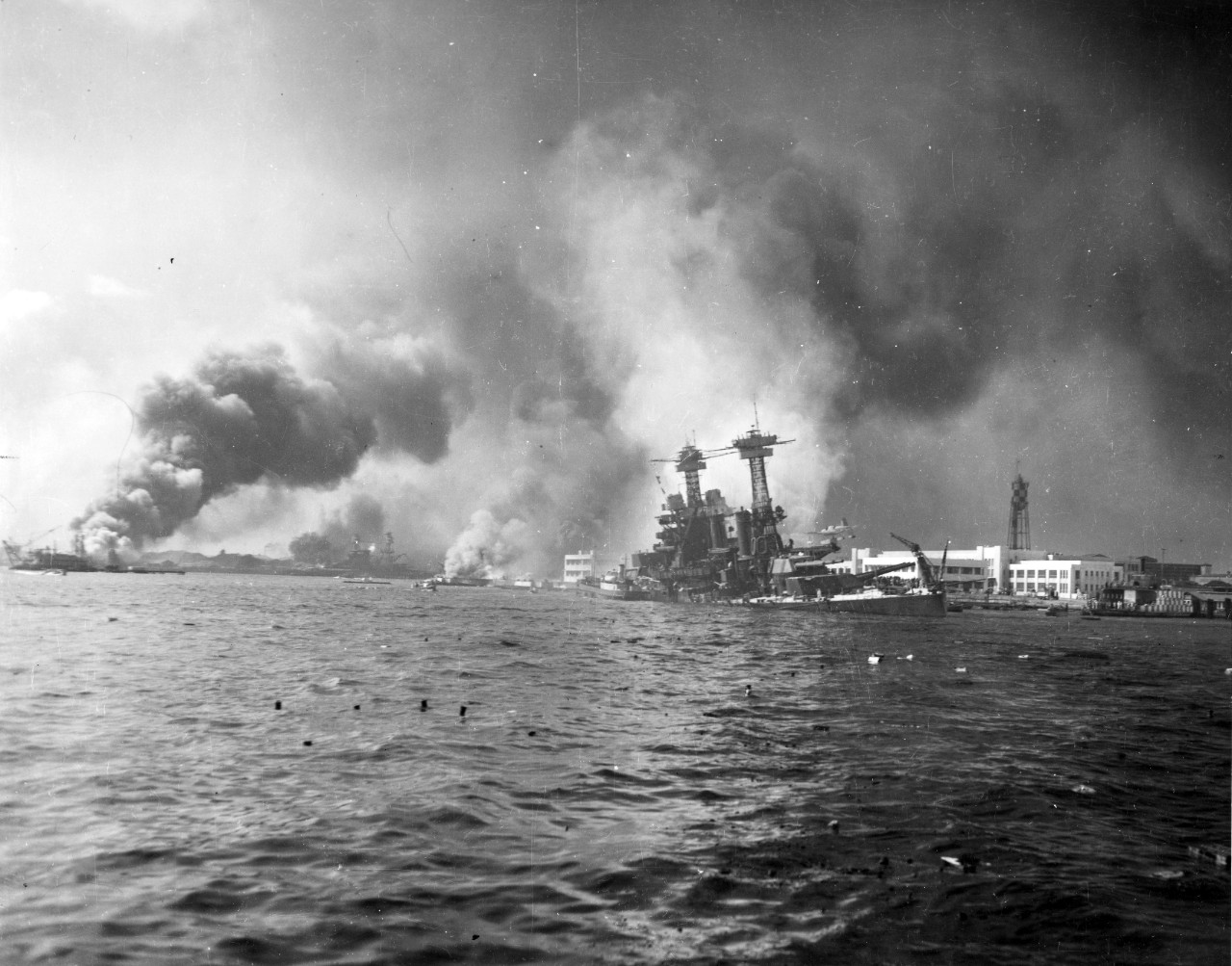 A view down Pearl Harbor’s main channel during the Japanese air attack, 7 Dec 1941. Note the battleship USS California listing and sinking and note the Ford Island water tower at right.