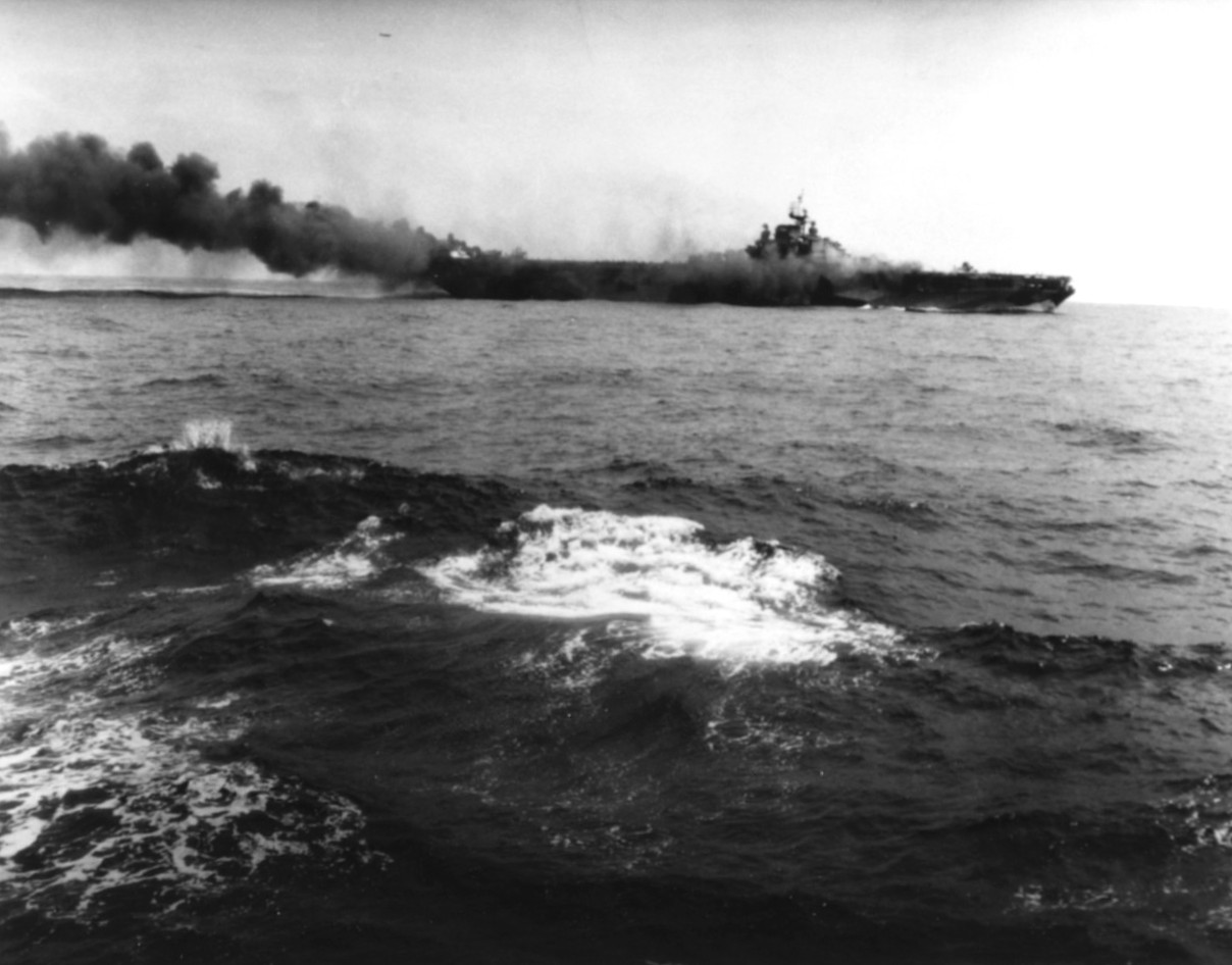 USS Hancock burning after being struck by a kamikaze special attack aircraft off Okinawa, 7 Apr 1945. Note fires burning fore and aft, and TBM Avenger flying over the carrier. Photographed from USS Pasadena.