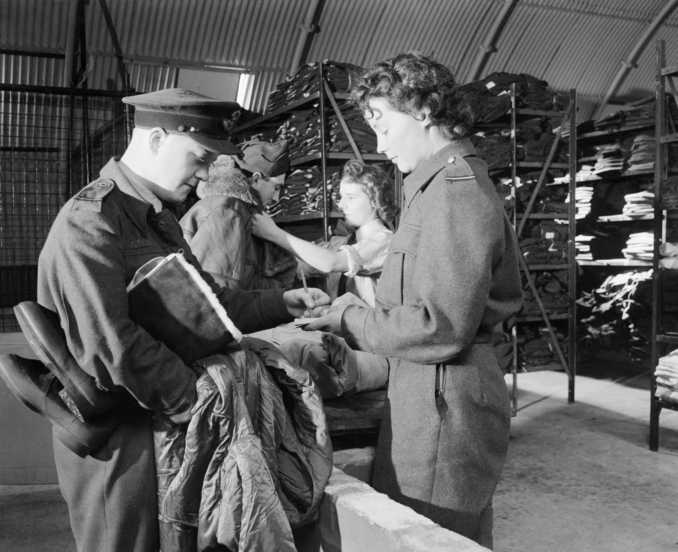 A WAAF section officer and her assistant issuing electrically-heated waist coat, 1940-pattern boots, and Irvin sheepskin flying jacket (background) to RAF bomber crewmembers, United Kingdom, Aug 1944