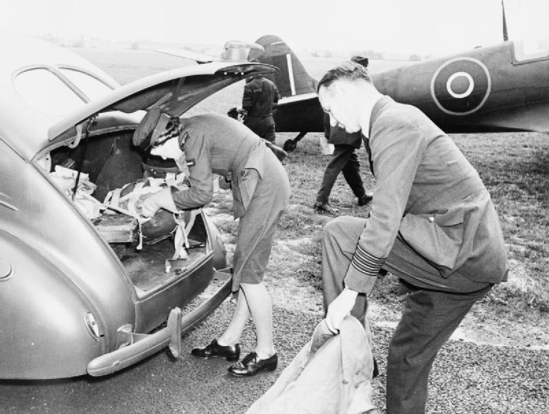 RAF Hornchurch station commander Group Captain Charles Lott putting on his flying kit with the assistance of his WAAF driver Leading Aircraftwoman Mary Ford, Hornchurch, Essex (now London Borough of Havering), England, United Kingdom, Jun 1942; note Spitfire fighter in background