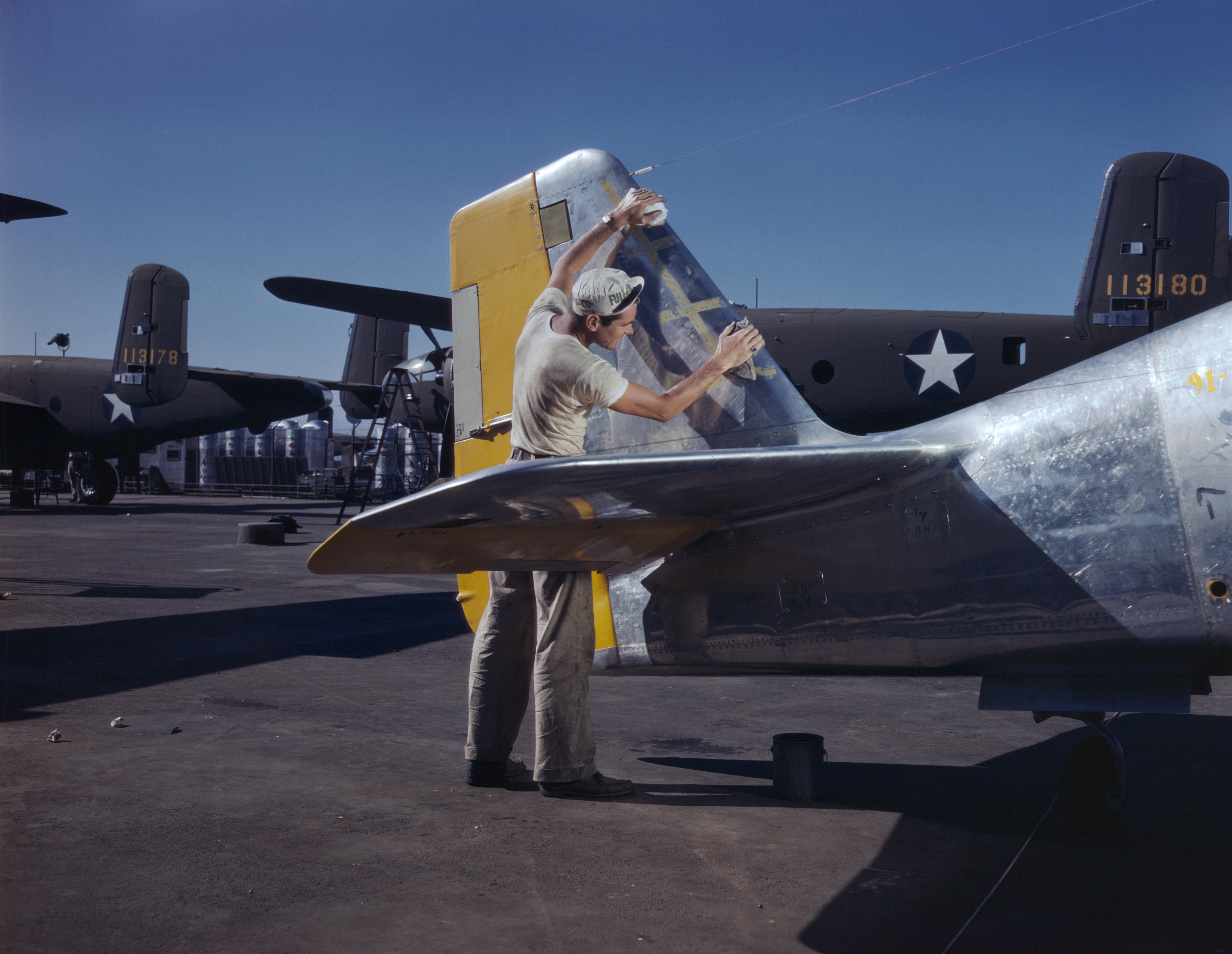 A North American Aviation worker preparing a P-51 Mustang fighter for painting, Inglewood, California, United States, Oct 1942; note B-25 Mitchell bombers in background