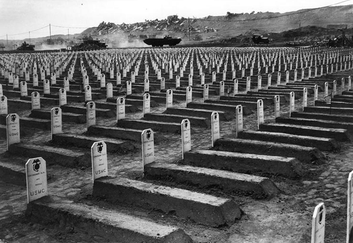 US 4th Marine Division cemetery on Iwo Jima, Japan, Mar 1945; note DUKW and jeep driving in background and wrecked Japanese aircraft beyond