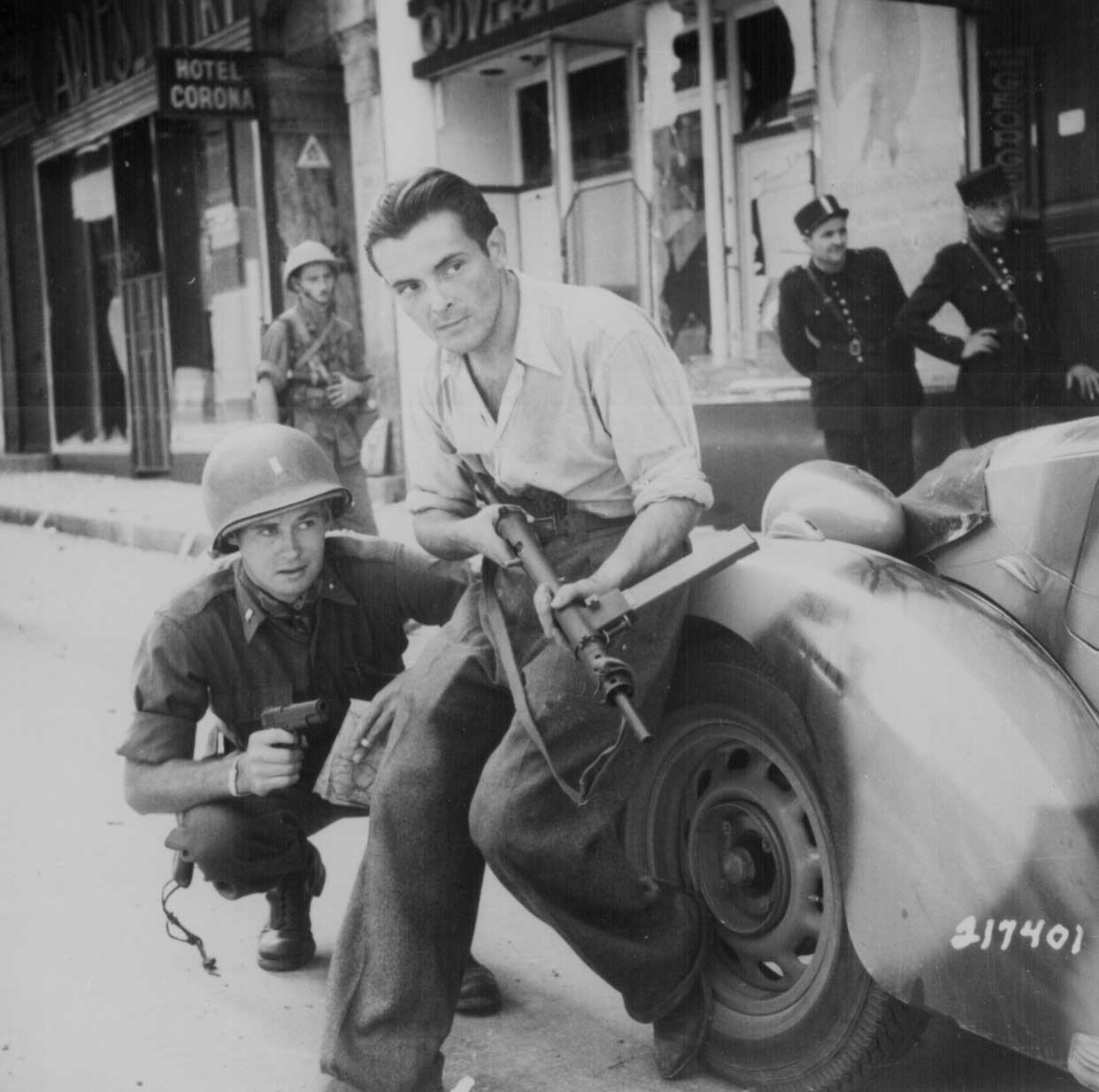 An American officer and a French partisan with a Sten sub-machinegun crouched behind a car during a street fight in a French city, Jun 1944