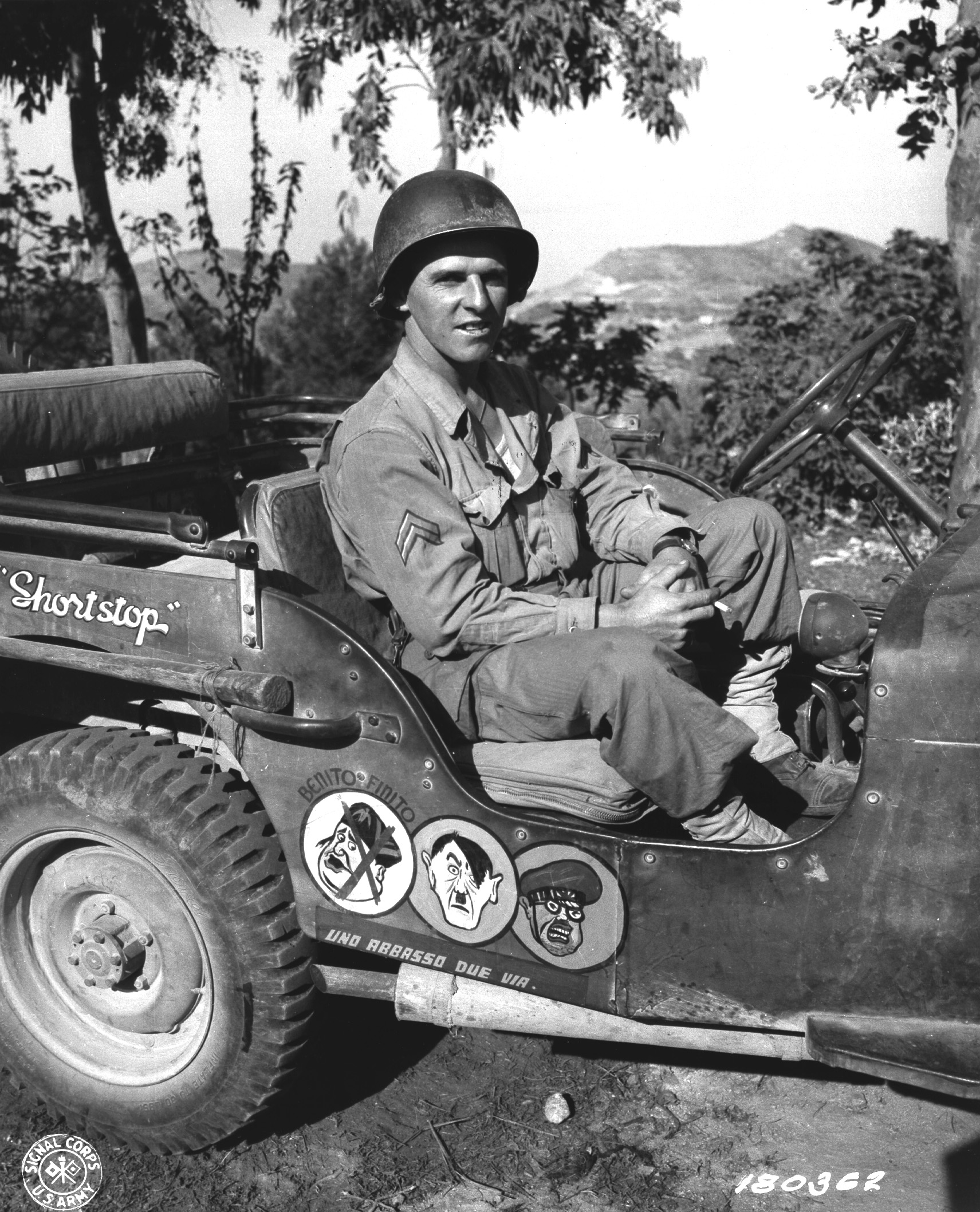US Army Corporal Paul F. Janesk posing in his jeep in Sicily, Italy, 3 Sep 1943; note cartoon of Axis leaders drawn on his jeep, and Mussolini crossed out