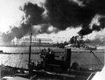 View of the Pearl Harbor drydocks from Ford Island during the Japanese air attack, 7 Dec 1941. Note seaplane tender USS Avocet in the foreground and battleship USS Nevada almost broadside to the channel.