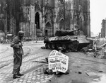 Corporal Luther E. Boger of US 82nd Airborne Division reading a warning sign in front of Cologne Cathedral, Köln (Cologne), Germany, 4 Apr 1945; note Thompson submachine gun and Panther tank wreck
