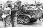 German officers speaking in front of a camouflaged SdKfz. 251 halftrack vehicle, Northern France, 21 Jun 1944; note German Army lieutenant with Iron Cross medal