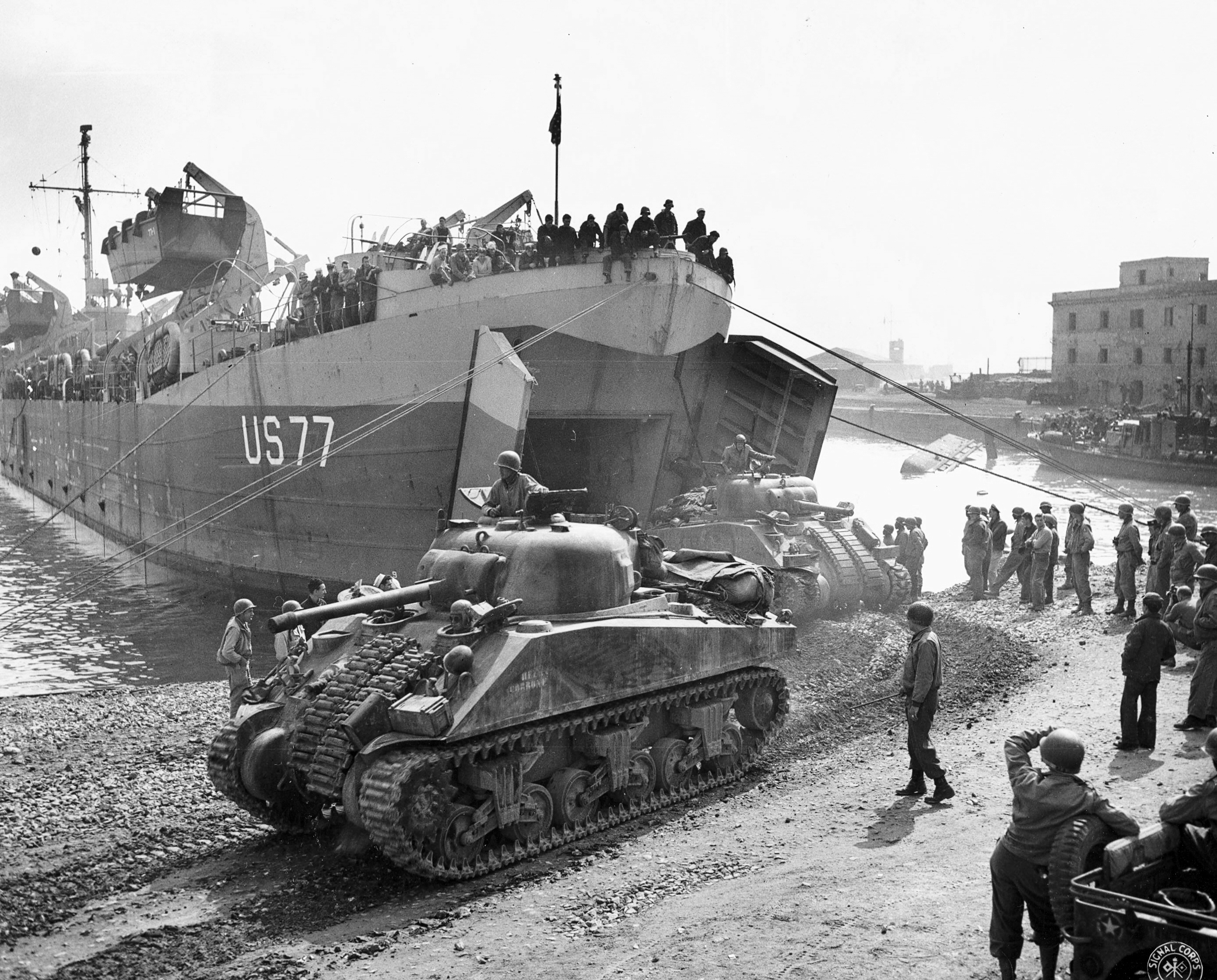 LST-77 off-loading M4 Sherman tanks at Anzio, Italy, May 1944; note the small barge capsized in the background and LCVP on the LST's davits